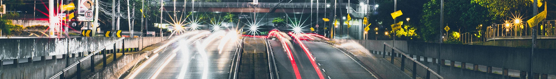 Long-exposure night shot of light trails on a busy urban roadway with street lights and signs.