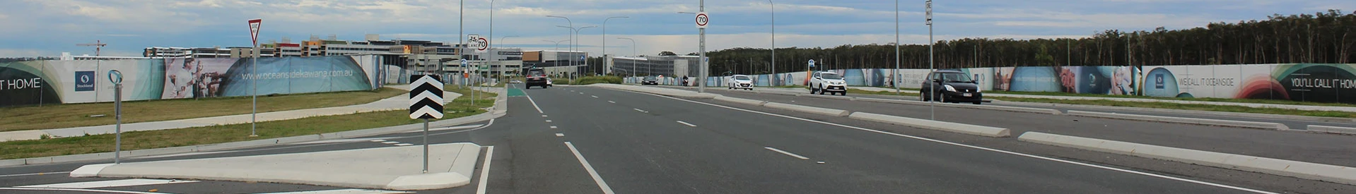 Wide road leading to a commercial area with advertising banners on the surrounding fences and several cars in view.